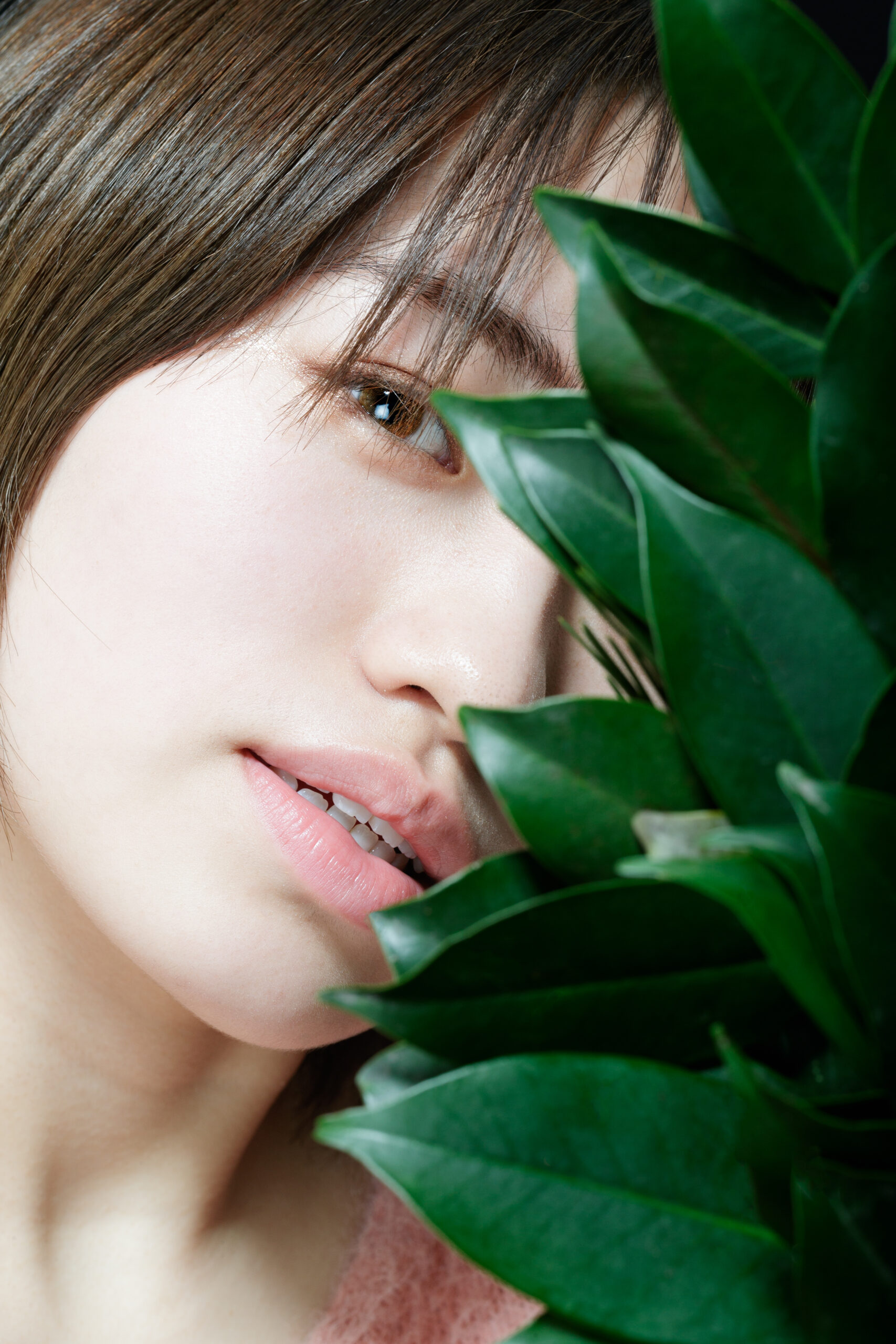 画像: 若い女性がグリーンの植物の葉越しに微笑むポートレート写真。ナチュラルメイクとソフトな色調が特徴。キーワード: 自然, 植物,