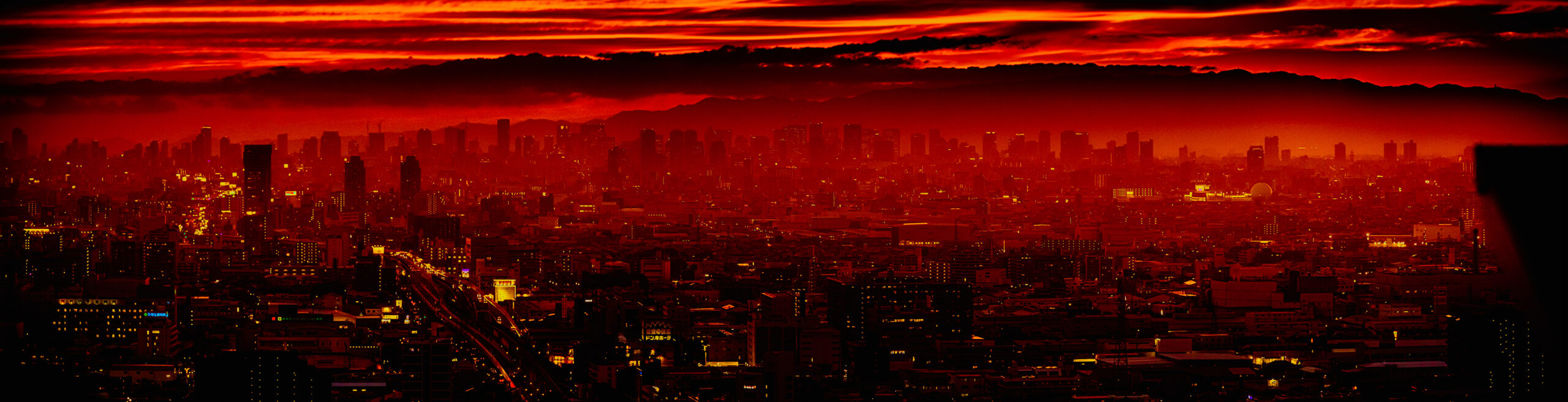 画像: 赤く染まる夕焼けの都市風景。シルエットの摩天楼が広がり、夜の訪れを感じさせる。夕焼け、都市、夜景、シルエットが