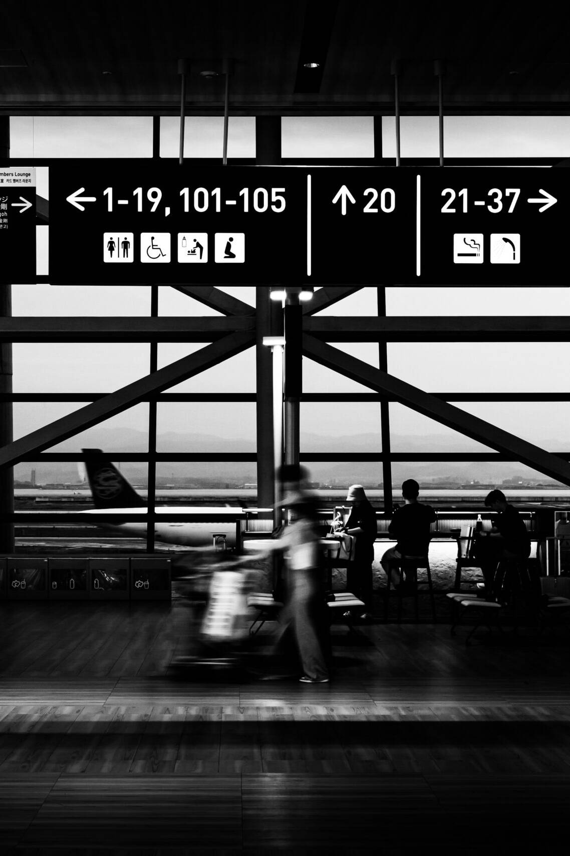 画像: モノクロ写真の空港ターミナル内で、案内標識と時計が高い天井から吊るされています。窓からの風景には滑走路が見え、乗客たちが座