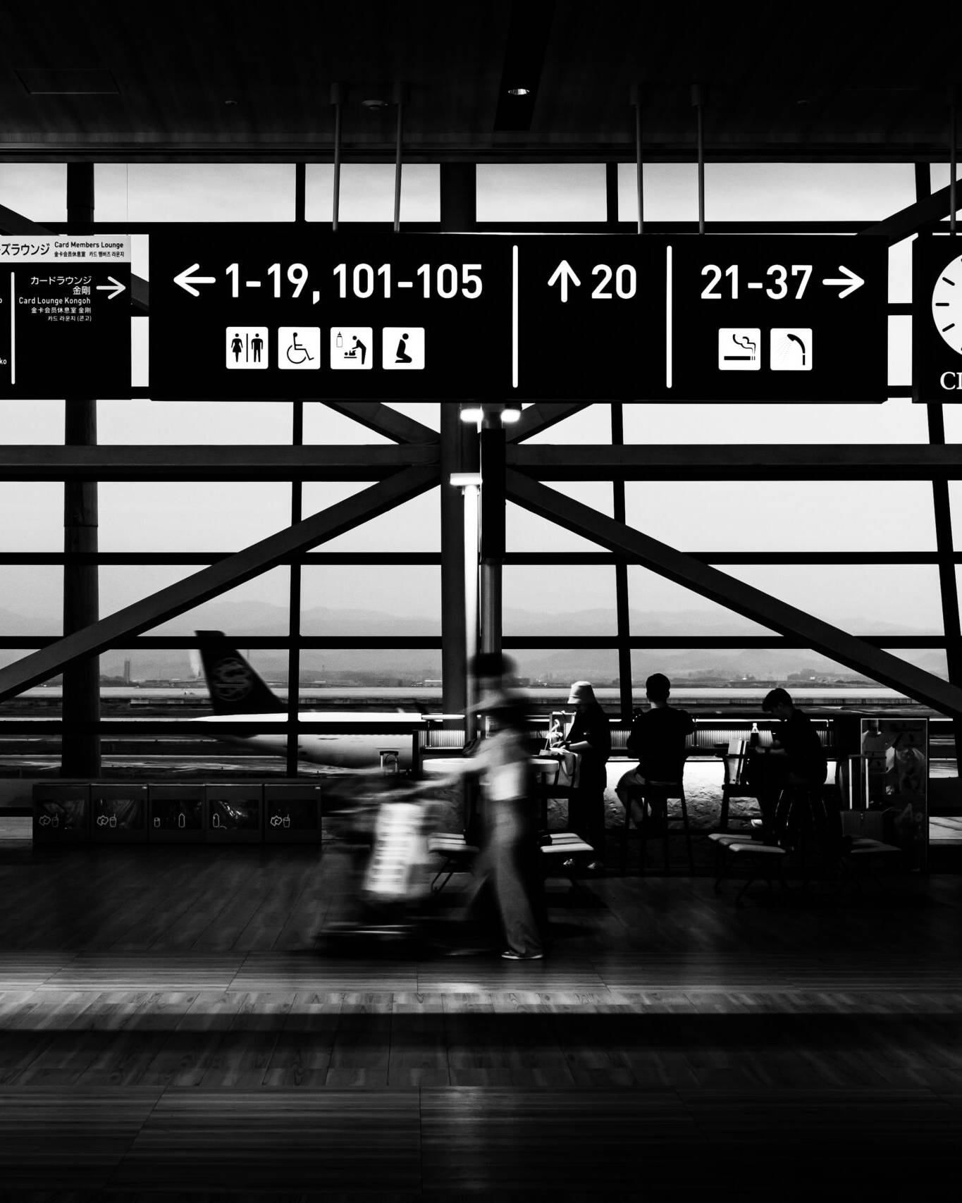画像: モノクロ写真の空港ターミナル内で、案内標識と時計が高い天井から吊るされています。窓からの風景には滑走路が見え、乗客たちが座