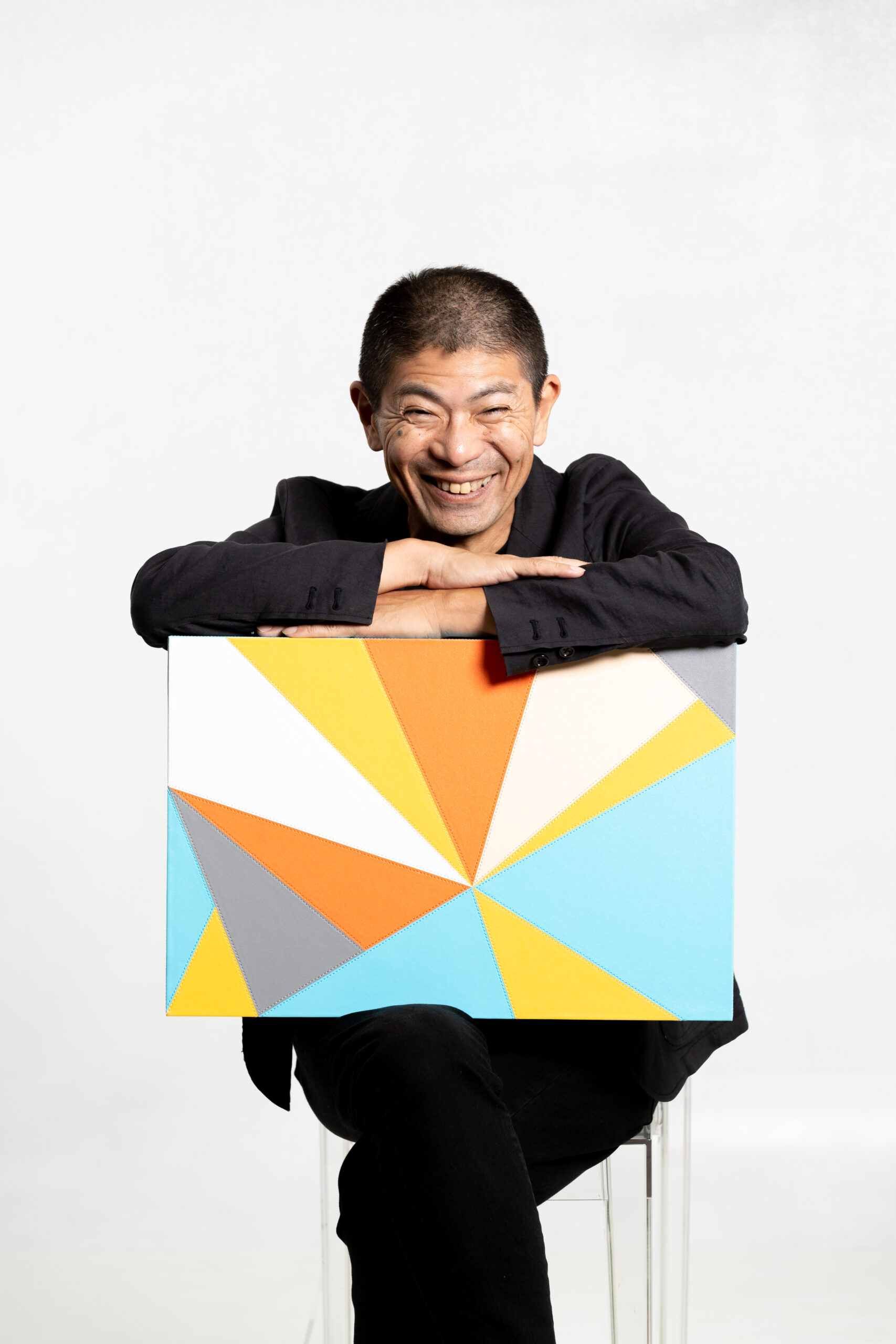 画像: 申し訳ありませんが、この画像の人物について特定することはできませんが、笑顔の男性が色鮮やかな幾何学模様のアートパネルを持っている写真です。白い背景が