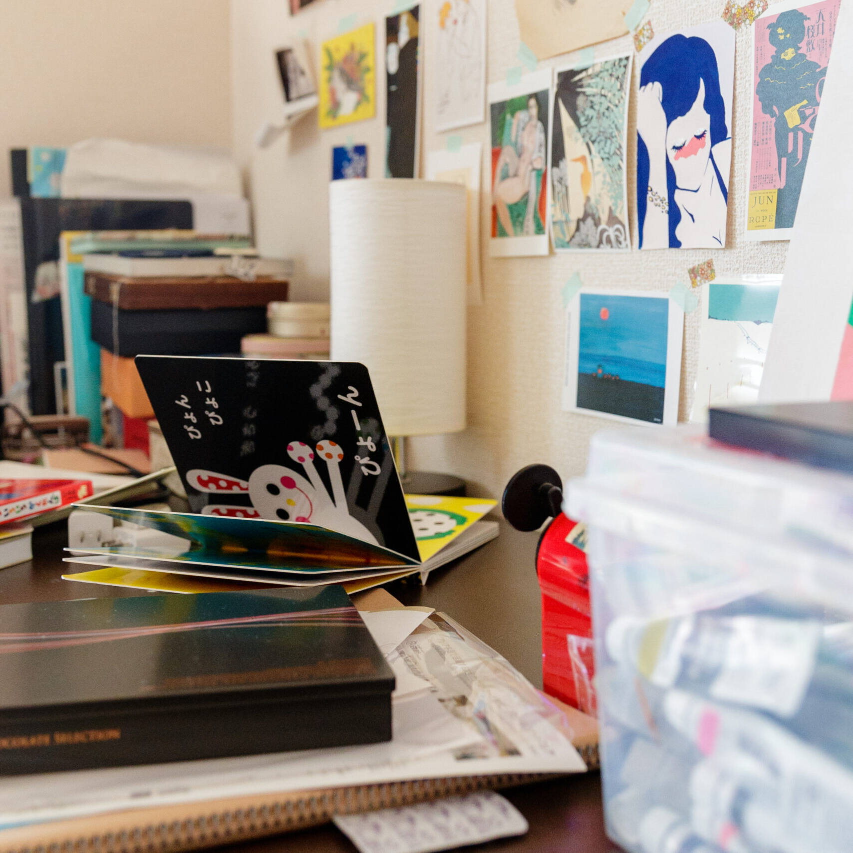 画像: 机の上には多様なアイテムが散らばっています。絵本、ペン、色彩豊かなボール、絵具があり、壁にはアート作品が飾られています。この場面