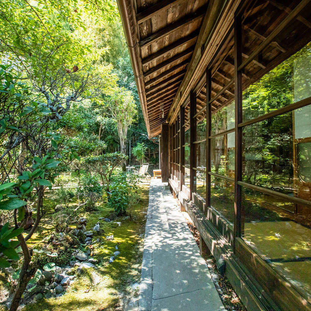 画像: 日本庭園の美しい景観を背景にした伝統的な和風建築の縁側です。豊かな緑に囲まれた静かな空間が広がり、自然との調和を感じ
