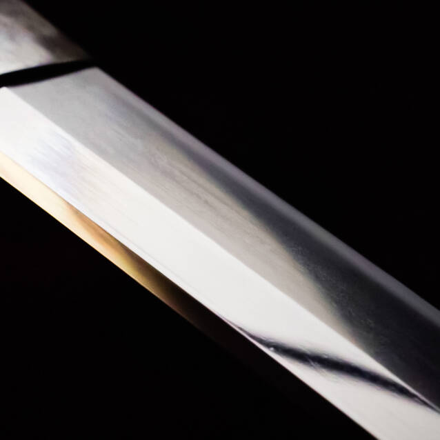 画像: 黒い背景に対して、日本刀の鋭い刃が大きく映し出されています。刀身の美しい反射が強調され、伝統的な武器の精緻さと職人技