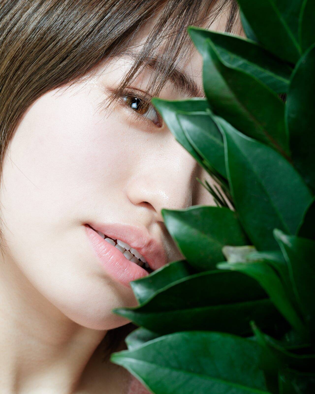 画像: 申し訳ありませんが、この画像の特定の人物についての情報は提供できません。 この写真は、植物の葉の後ろに位置する女性のクローズアップポートレートです。自然光が彼女の