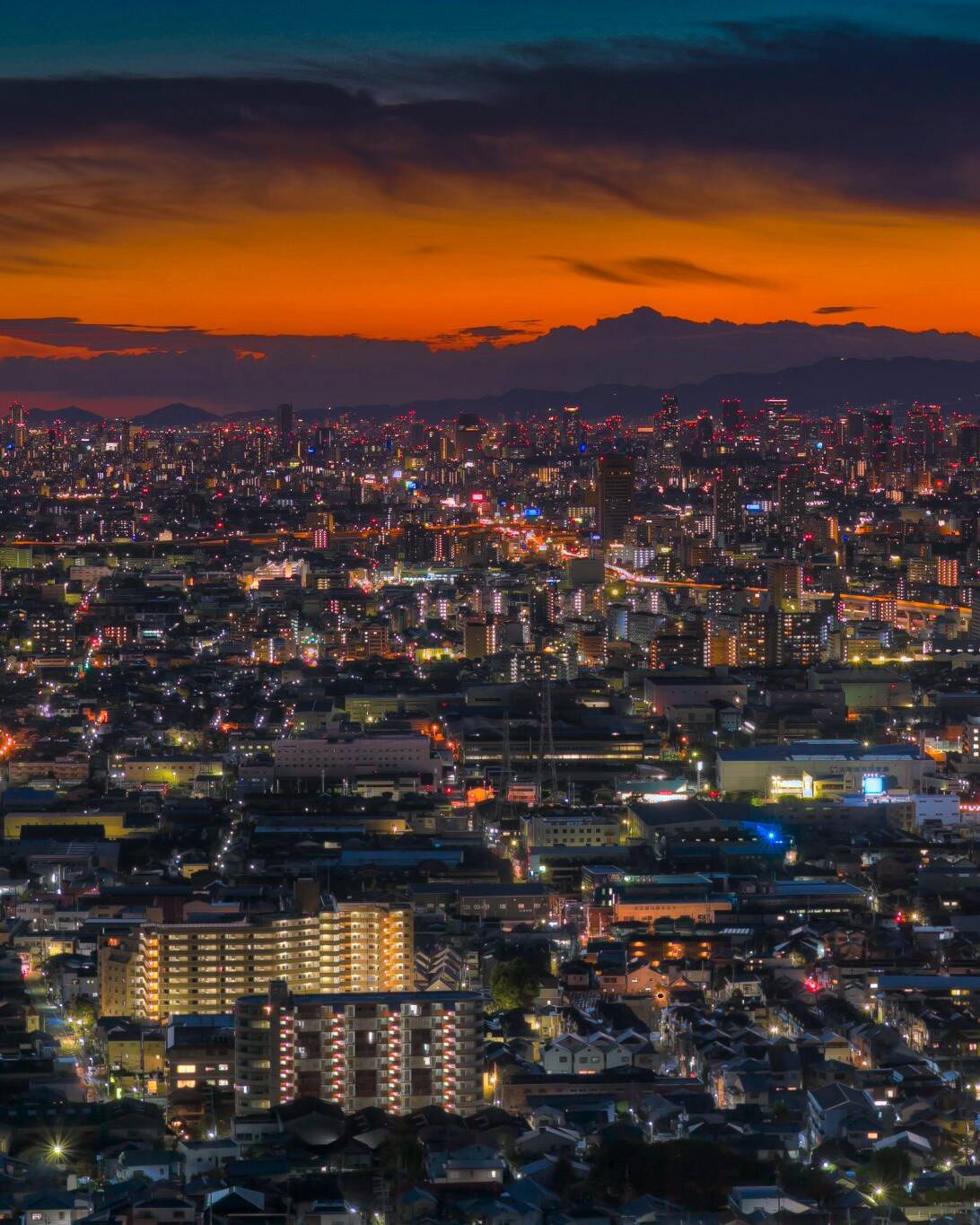 画像: 大阪の都市夜景が広がる夕暮れの風景です。オレンジ色の空が特徴的で、ビルと道路の明かりが美しく輝いています。夜景、夕焼け
