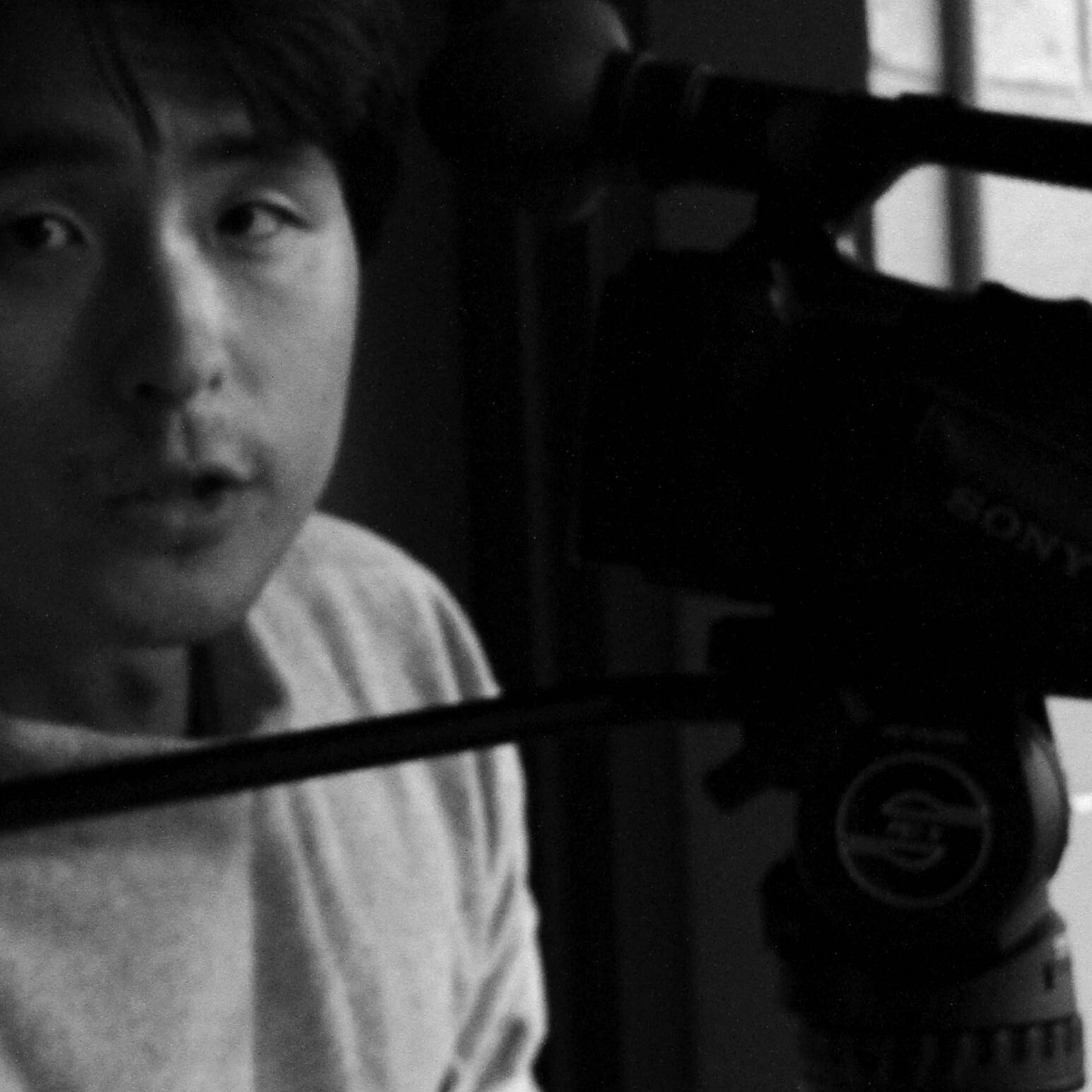 画像: 白黒の写真に映る男性が、カメラのそばで真面目な表情をしています。撮影や映画制作、ドキュメンタリーのシーンを連想させる。カメ