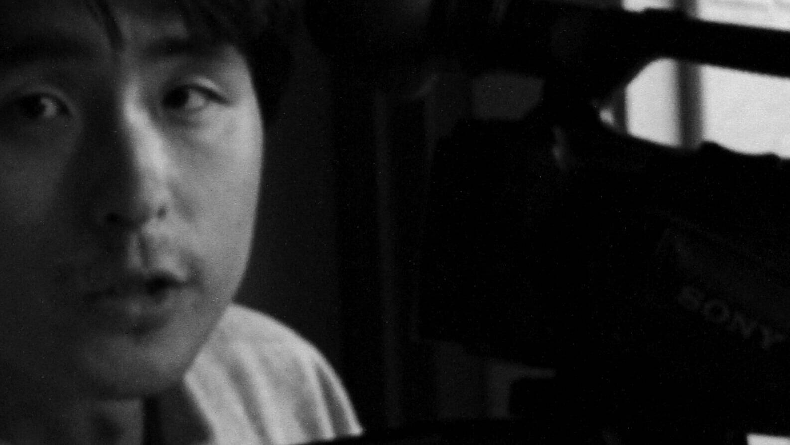 画像: 申し訳ありませんが、この画像から特定の個人を識別することはできません。画像には、ビデオカメラを操作している人物が写っています。モノクロ写真で、カメラには「