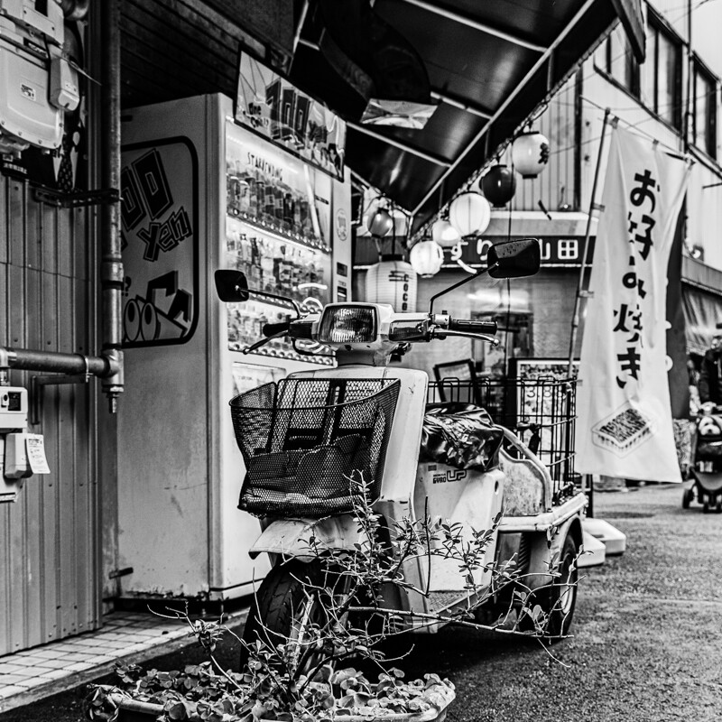 画像: 白黒の画像には、レトロな雰囲気のスクーターが路地に停められています。背景には自動販売機と「お好み焼き」の旗、提灯があり、昭