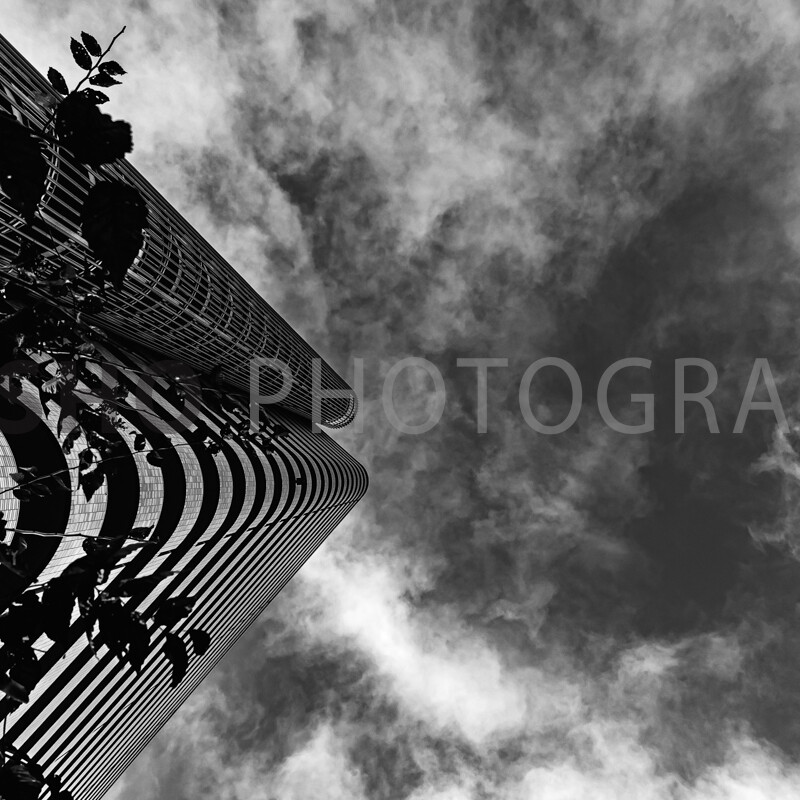 画像: 高層ビルの外観とドラマチックな雲が白黒で捉えられた写真です。視点を下から上に向け、ビルの曲線的なデザインと空