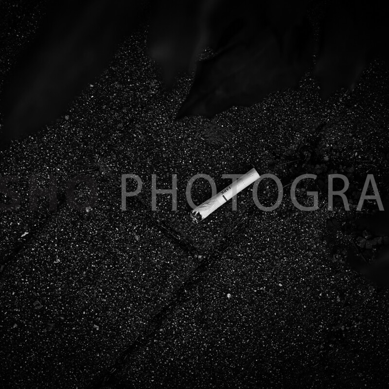 画像: 白黒写真にて、アスファルトの地面に落ちた煙草の吸い殻が映されています。夜の都市風景を彷彿とさせるシーンで、路上のゴ