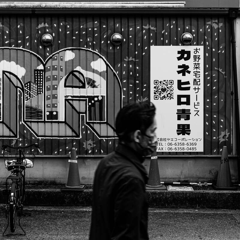 画像: この白黒写真は、都市のストリートビューを捉えており、壁には看板とアートが描かれています。看板には「カネヒロ青果」と書かれた野菜宅