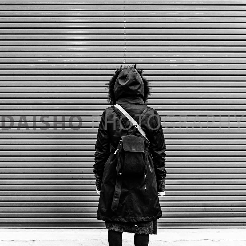 画像: 黒いコートを着た人物が背面からシャッターの前に立っている白黒写真。都市のストリートファッション、匿名性、後ろ姿が際立つシーン。 キーワード: 白
