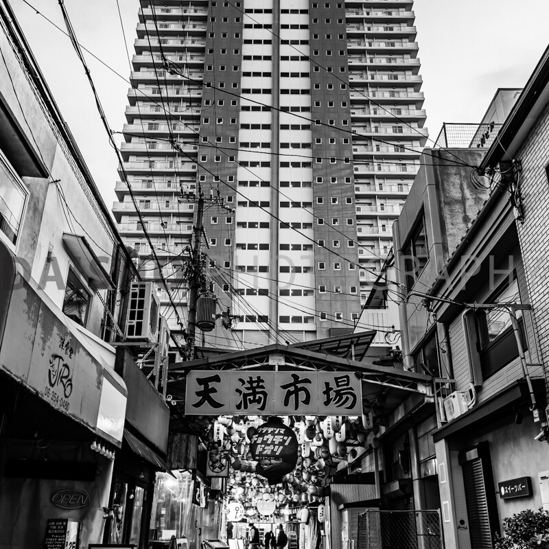 画像: 都市のアーケード入り口の写真です。天満市場の看板がポールに掲げられ、背景には高層マンションがそびえ立っています。細い路地に位置する市場は