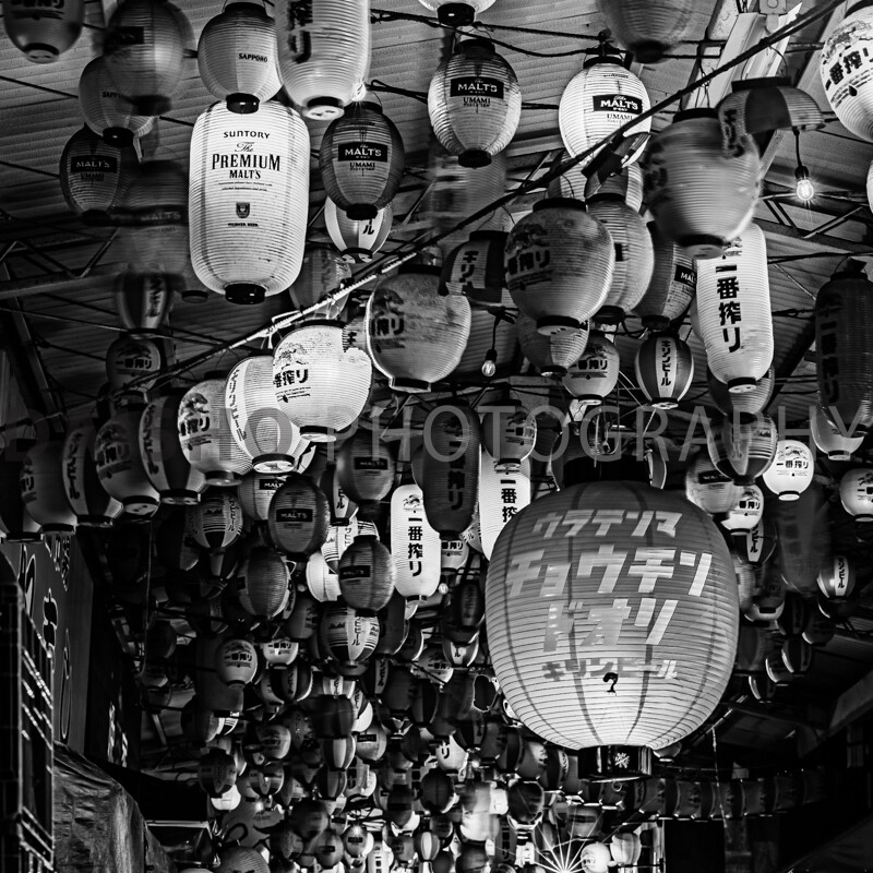 画像: 日本の商店街で撮影された白黒写真で、たくさんの提灯が軒先に吊るされています。夜の雰囲気が漂うこのシーンは、伝統的な日本の文化