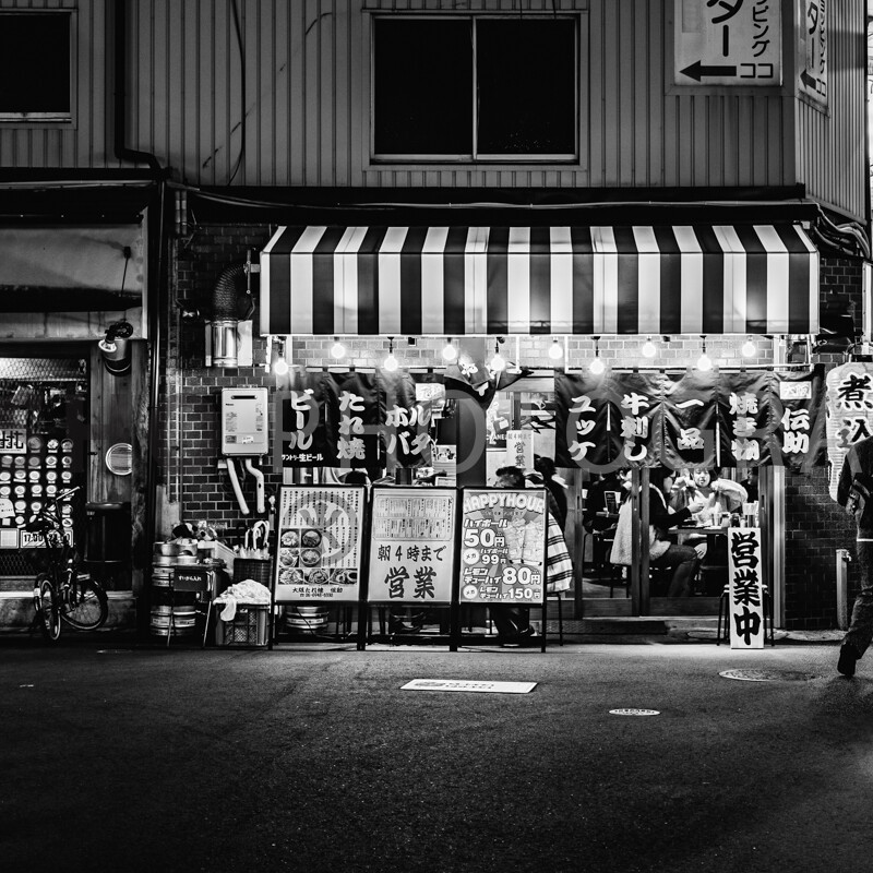 画像: この画像は、日本の繁華街にある居酒屋の夜景をモノクロで描写しています。居酒屋の店頭には様々なメニューが掲示され、通りには人々が行き