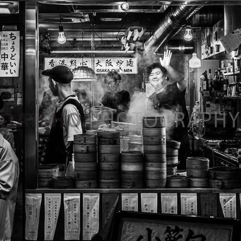 画像: 大阪の飲食店で働くスタッフが手を振るシーンが白黒で写されています。店内は蒸気が立ち込め、多くのせいろが並んでおり、活気があります