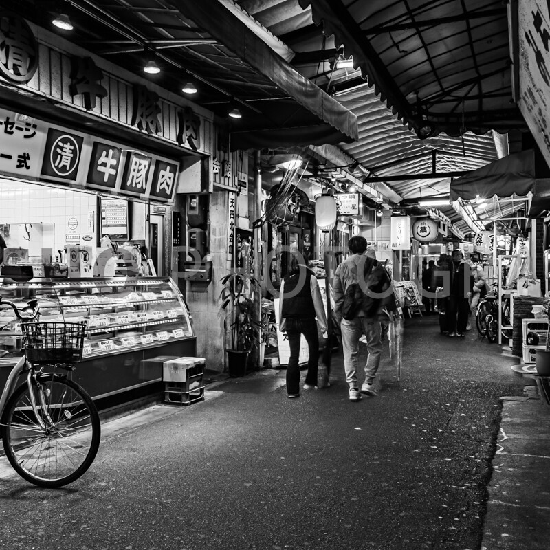 画像: これは、アーケード商店街の写真で、白黒で撮影されています。肉屋の商品がディスプレイに並べられており、自転車が店の前に置かれています。人々