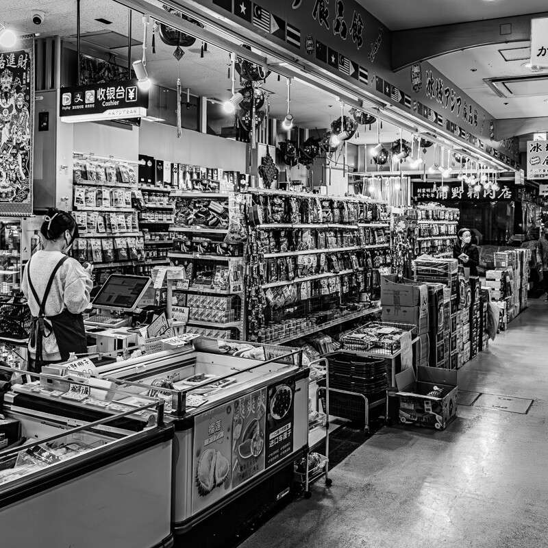 画像: 日本のアジア系スーパーマーケットの店内のモノクロ写真。商品棚には多様な食品が整然と並び、エプロンを着た店員がレジで接客をしています