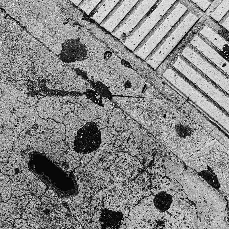 画像: 白黒の画像には、ひび割れたアスファルトと線状のコンクリートブロックが交差しています。雨跡が点在しており、都市の歩道のディテールが