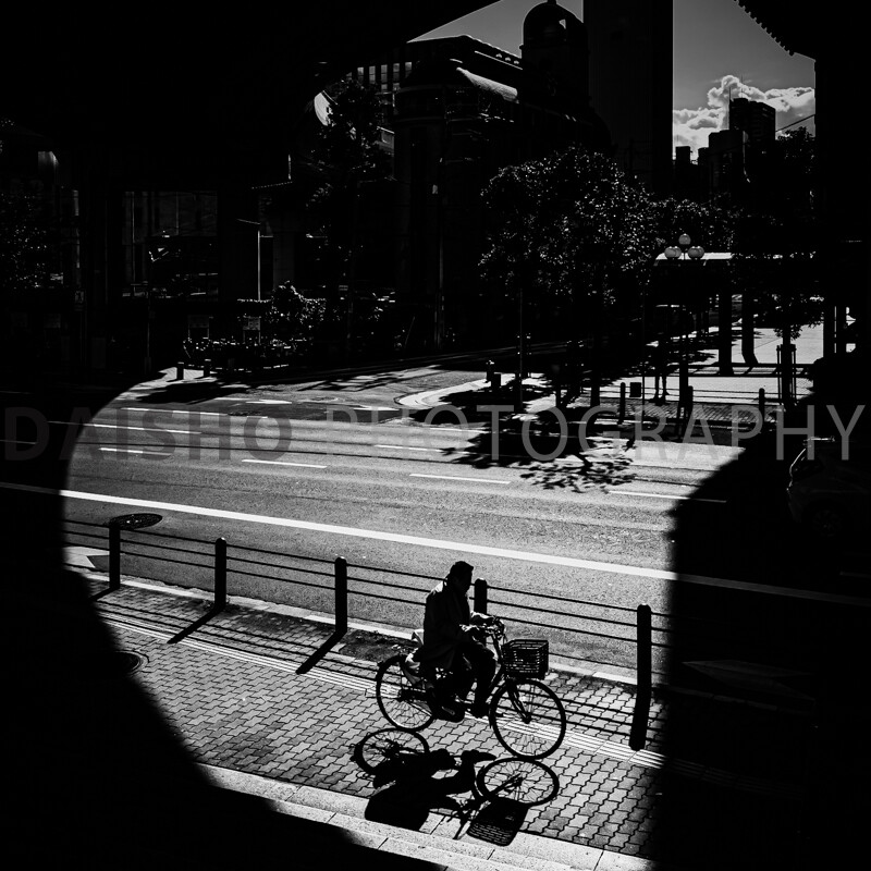 画像: 黒白の都市風景で、自転車に乗る人が影の中を走行しています。強いコントラストが印象的なストリートフォトグラフィーで、光と影が交