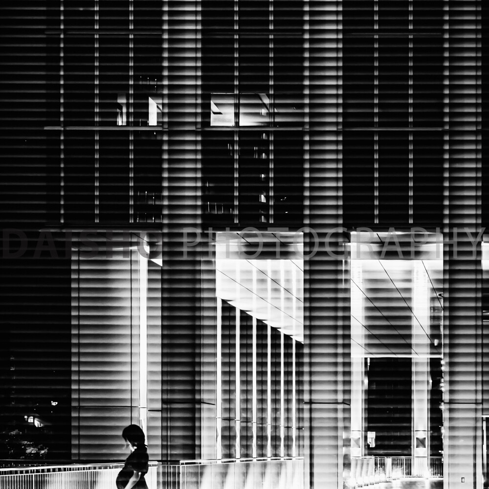画像: ビルの外壁と通路を背景に、夜間に照明が美しく映えるモノクロの現代的な都市風景です。人が歩道に立っており、光と影のコ