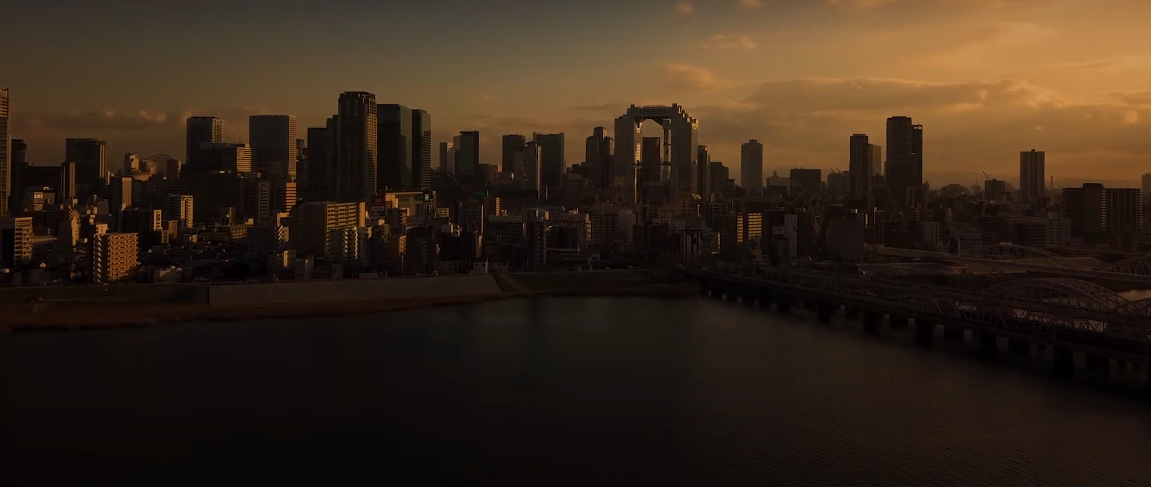 画像: 大阪市の夕暮れ時のスカイライン、建物や高層ビルが温かい夕日の光に照らされて美しいシルエットを形成。淀川も見える。都市